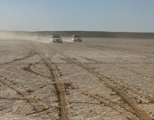 Un groupe de véhicules soulevant de la poussière alors qu'ils traversent un paysage désertique plat et étendu, avec des traces de pneus s'entrecroisant au premier plan.