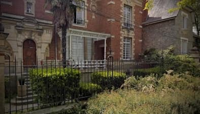 Maison de ville en briques rouges avec une entrée surmontée d'un auvent en verre et une porte en bois, entourée d'une clôture noire et d'une végétation abondante.