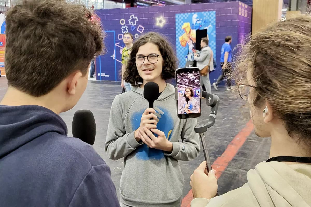 Un jeune homme tenant un micro est interviewé dans un espace public. Une autre personne filme la scène avec un smartphone monté sur un stabilisateur, tandis qu'un troisième individu lui pose des questions, micro en main. L'écran du téléphone montre le visage de l'interviewé en gros plan. En arrière-plan, on aperçoit des décorations colorées sur les murs, avec des motifs géométriques et des personnages, créant une ambiance vivante et dynamique. La scène semble se dérouler lors d'un événement ou dans un lieu dédié aux interactions médiatiques.