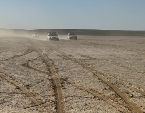 Un groupe de véhicules soulevant de la poussière alors qu'ils traversent un paysage désertique plat et étendu, avec des traces de pneus s'entrecroisant au premier plan.