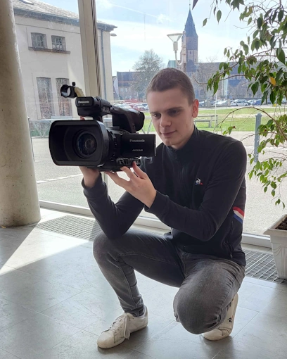Un homme est accroupi tenant une caméra vidéo professionnelle. Il est concentré sur le cadrage, regardant à travers le viseur de l'appareil. Des plantes d'intérieur et des reflets de lumière ajoutent à l'ambiance sereine de la photo.