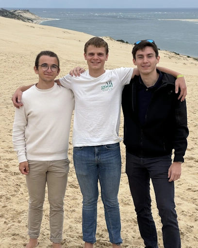 Trois hommes posent ensemble sur une dune de sable, en bord de mer. Ils se tiennent par les épaules avec une expression détendue. En arrière-plan, on peut apercevoir une étendue d'eau et des dunes qui s'étendent à perte de vue. Le ciel est nuageux, donnant à la scène un aspect calme et naturel.