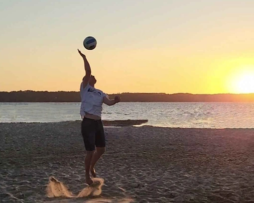 Un joueur de volley-ball est en plein service sur une plage au coucher du soleil. La balle est en l'air alors qu'il tend son bras pour la frapper. Le sable s'envole légèrement sous ses pieds. En arrière-plan, on aperçoit l'eau calme et le soleil descendant derrière l'horizon, illuminant la scène d'une lumière dorée. L'ambiance est paisible, avec un ciel clair qui commence à prendre des teintes chaudes.