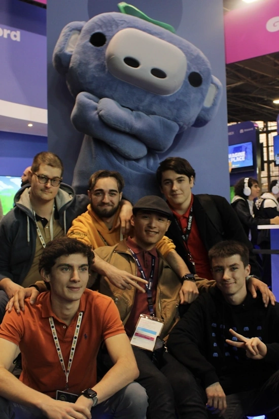 Des membres de l'agence FCA posant pour une photo avec une grande mascotte du personnage Wumpus de Discord en arrière-plan. Ils sont à un événement, identifiable par les badges et les cordons autour de leurs cous et l'ambiance générale de l'espace d'exposition avec des logos de Discord et de la publicité colorée en arrière-plan.